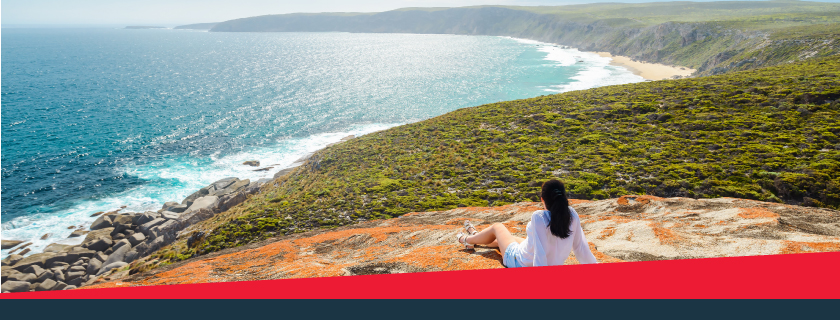 Woman reclining on rocky outcrop. She is looking out toward the ocean on her left side and there are green fields along the coast to her right.